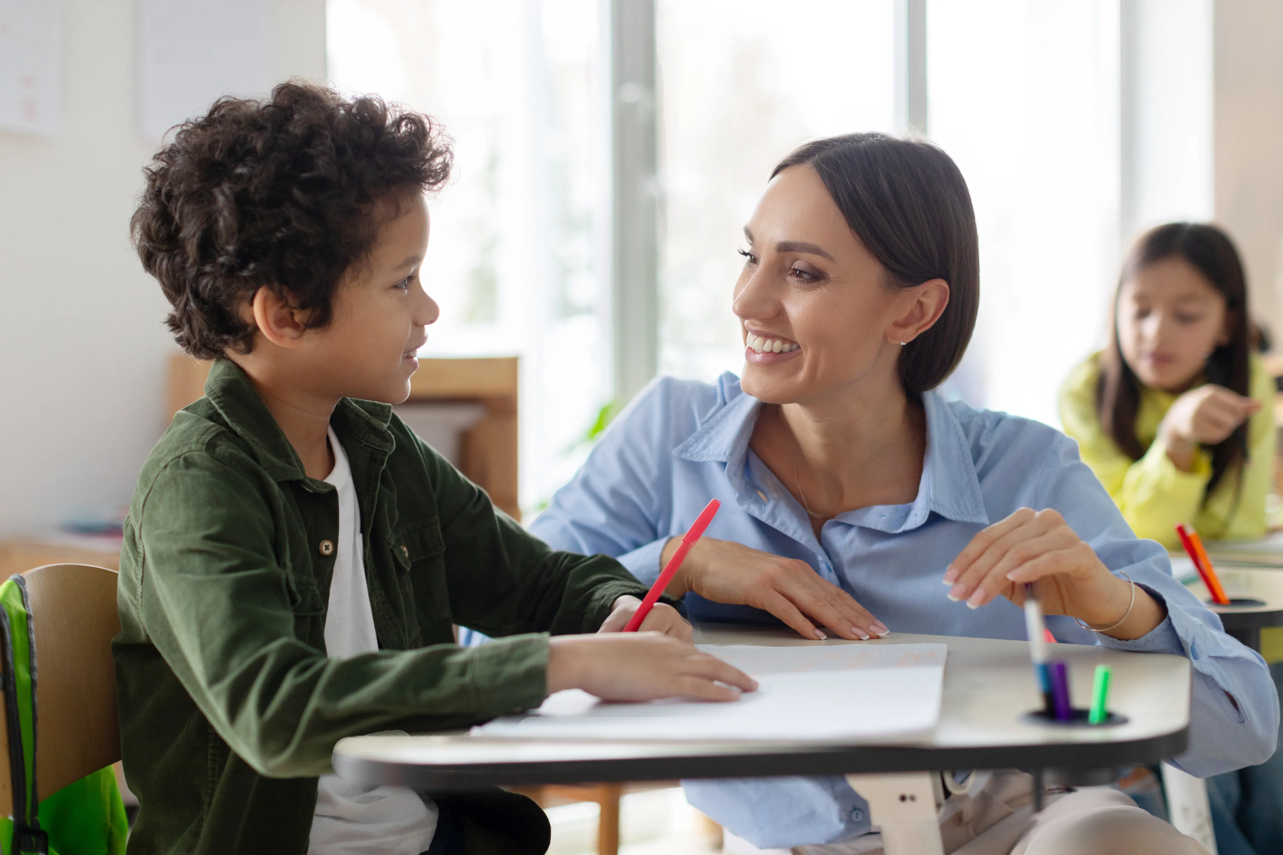 A teacher and student smiling together at a classroom table with markers and paper